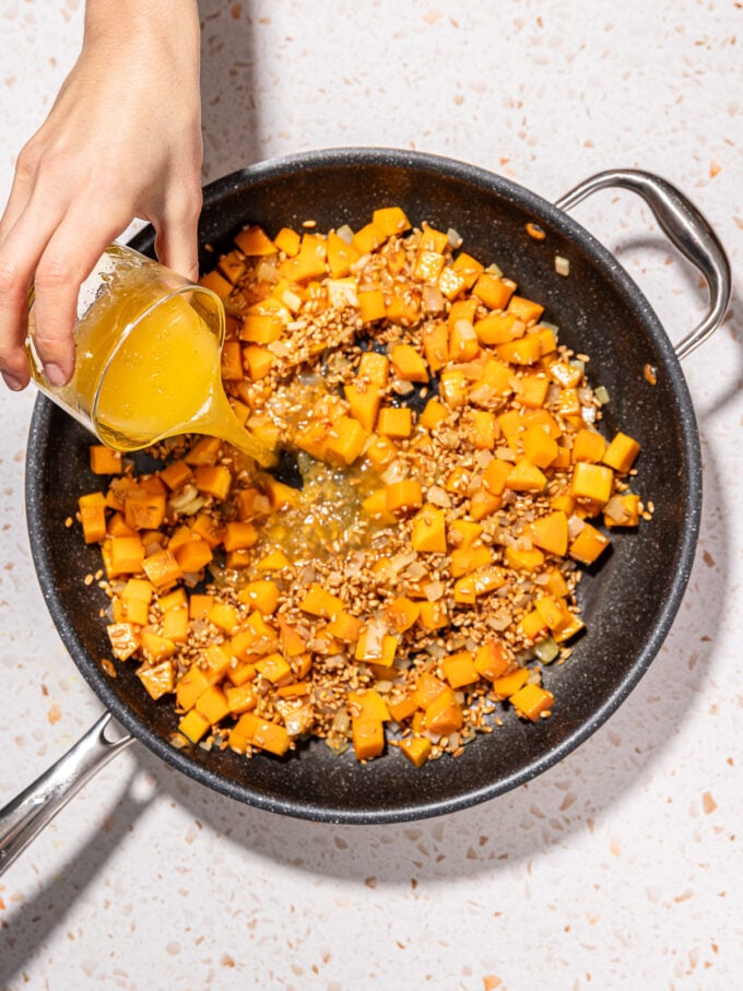 hand pouring broth into skillet with farro and squash