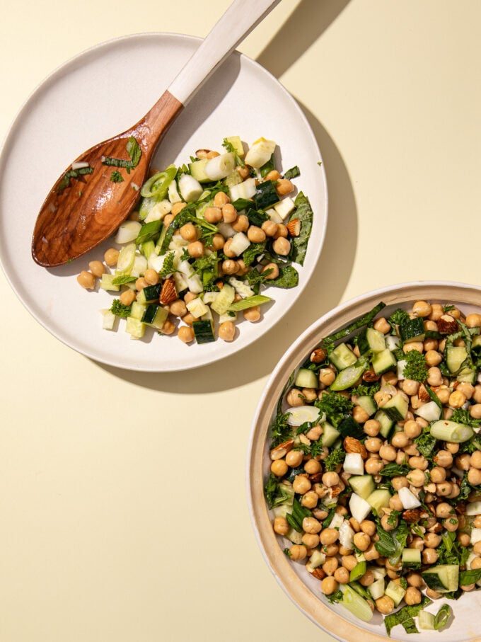 Chickpea cucumber salad on white plate with wooden spoon and bowl on yellow background