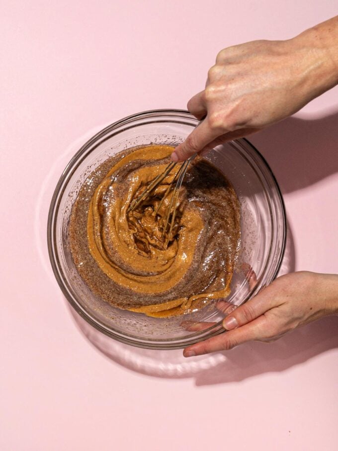 hand stirring peanut butter and maple syrup in glass bowl on pink background
