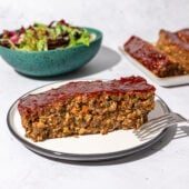 slice of lentil walnut loaf on white plate with blue bowl of salad behind it