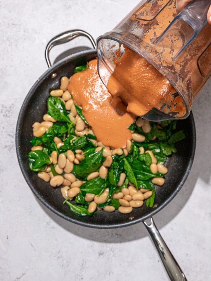 pouring rose sauce out of a blender onto a skillet with butter beans and spinach