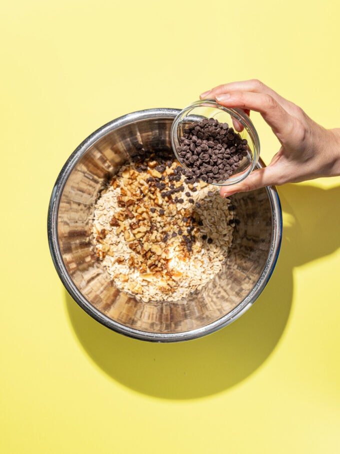 hand pouring chocolate chips into bowl of oats and walnuts