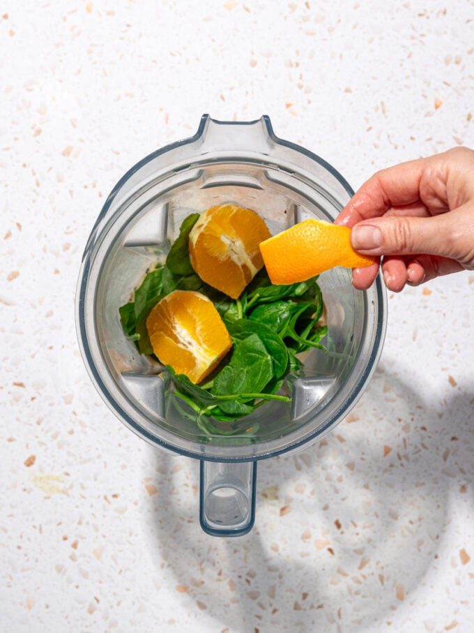 hand placing orange peel into blender with spinach and orange