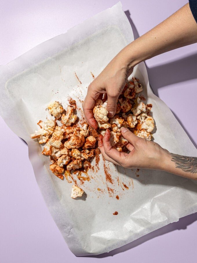 hand tossing cauliflower with spices on parchment lined baking sheet