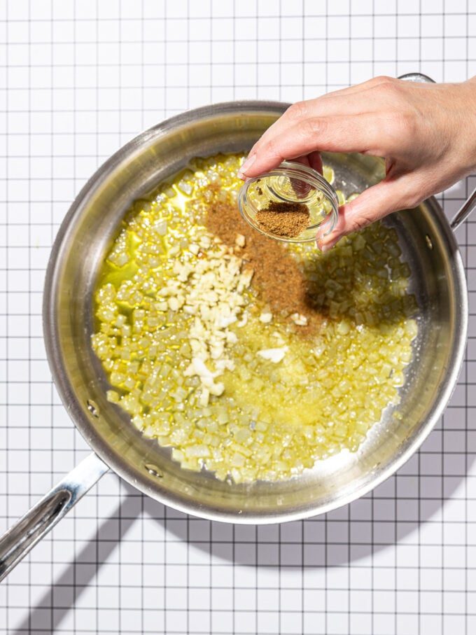 hand pouring cumin and garlic into cooked onions in skillet