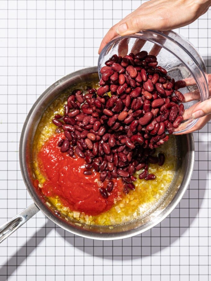 hand pouring bowl of kidney beans into skillet with tomato sauce and onions
