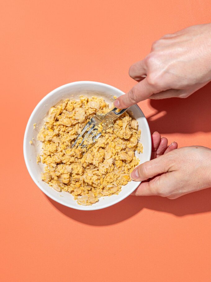 hand smashing chickpeas in white bowl with fork