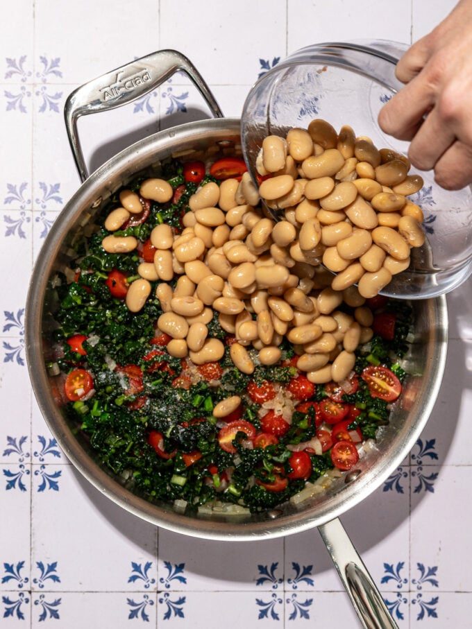 hand adding white beans to skillet with kale and tomatoes