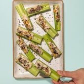 hand holding tahini stuffed celery stick next to rectangular plate of stuffed celery with seeds on top