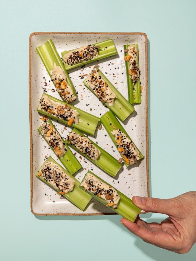 hand holding tahini stuffed celery stick next to rectangular plate of stuffed celery with seeds on top