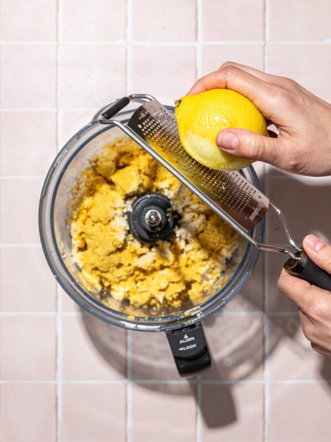 hand grating lemon zest over food processor with tofu and nutritional yeast