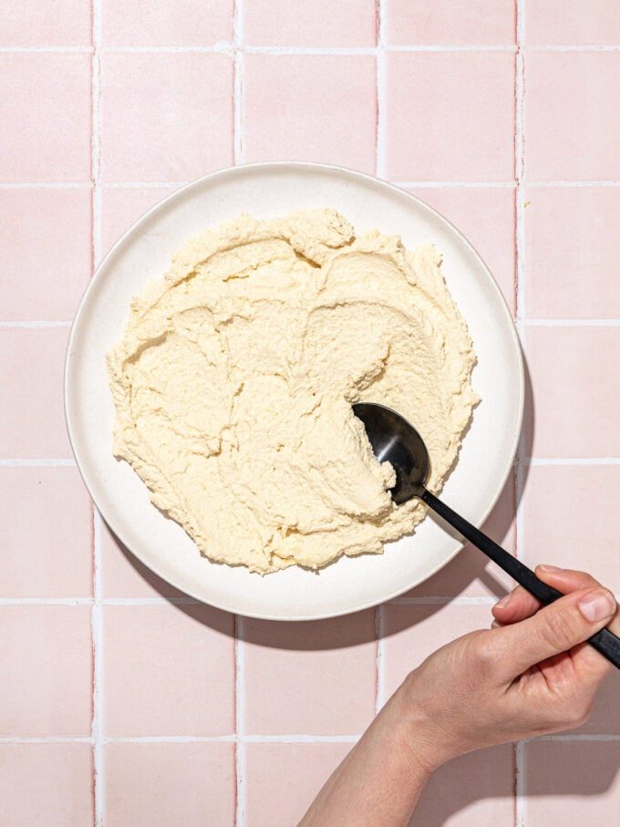 hand scooping tofu ricotta on white plate with black spoon on pink tile background