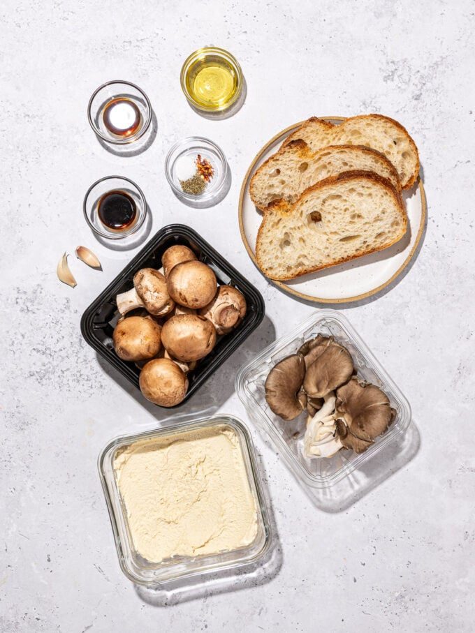 containers of mushrooms, tofu ricotta in a glass dish, slices of bread, bowls of oil, spices and soy sauce