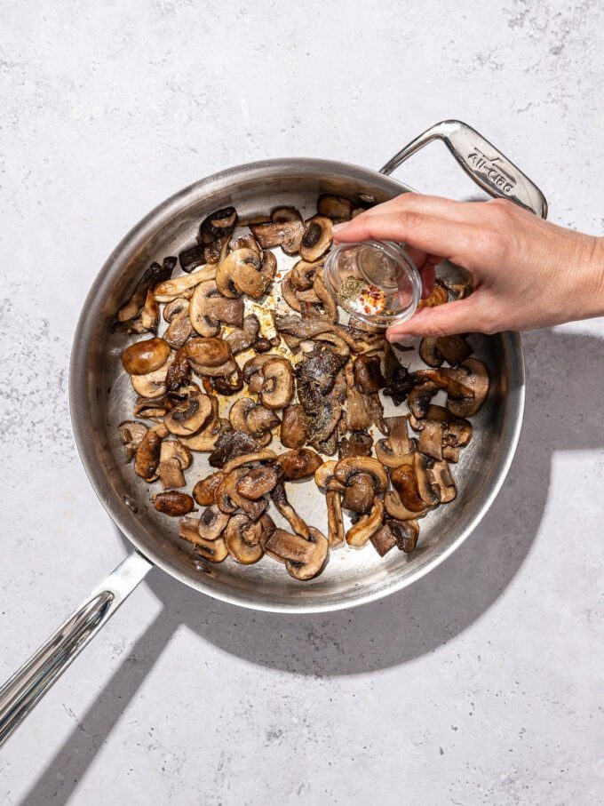 hand sprinkling spices into steel skillet with mushrooms