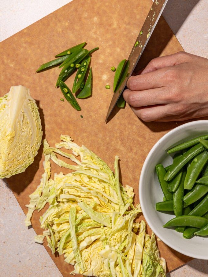 hand slicing snap peas on cutting board with shredded cabbage and bowl of snap peas