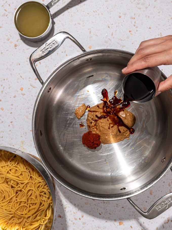 hand pouring soy sauce out of measuring cup into pot with peanut butter and curry paste