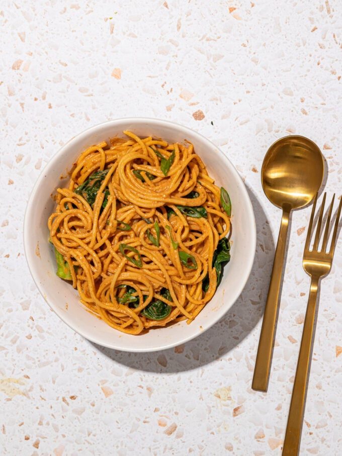 white bowl with peanut butter noodles and greens with a copper spoon and fork beside it