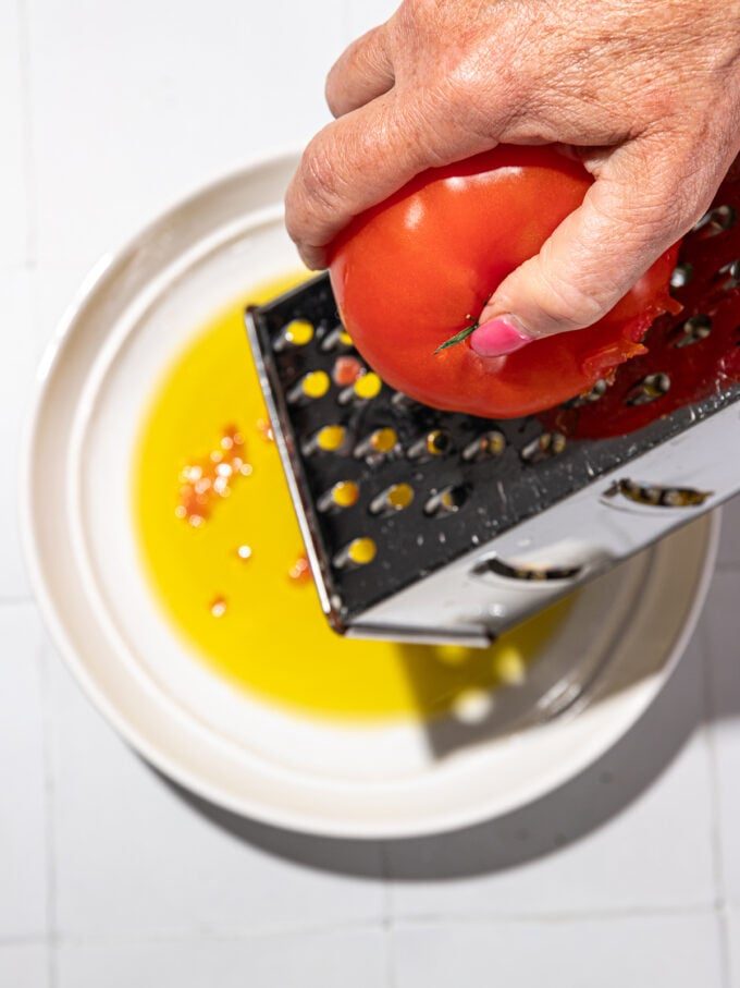hand grating tomato on box grater positioned over plate with olive oil