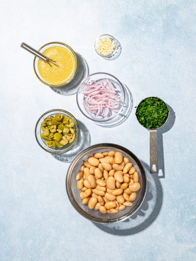glass bowl of white beans beside bowls of green olives, sliced red pepper, lemon dijon dressing and parsley