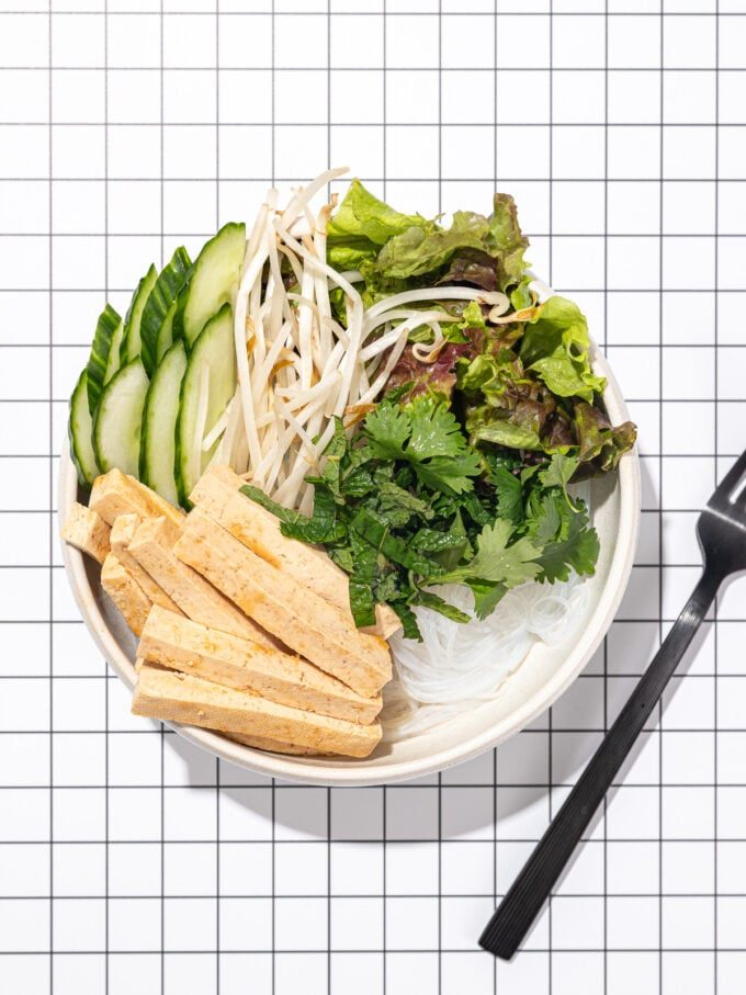 tofu, vermicelli noodles, lettuce and herbs in a white bowl with a black fork laying beside it