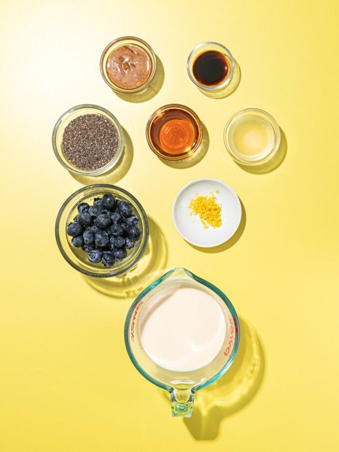 bowls of ingredients on a yellow background: soy milk, blueberries, maple syrup, chia seeds, almond butter