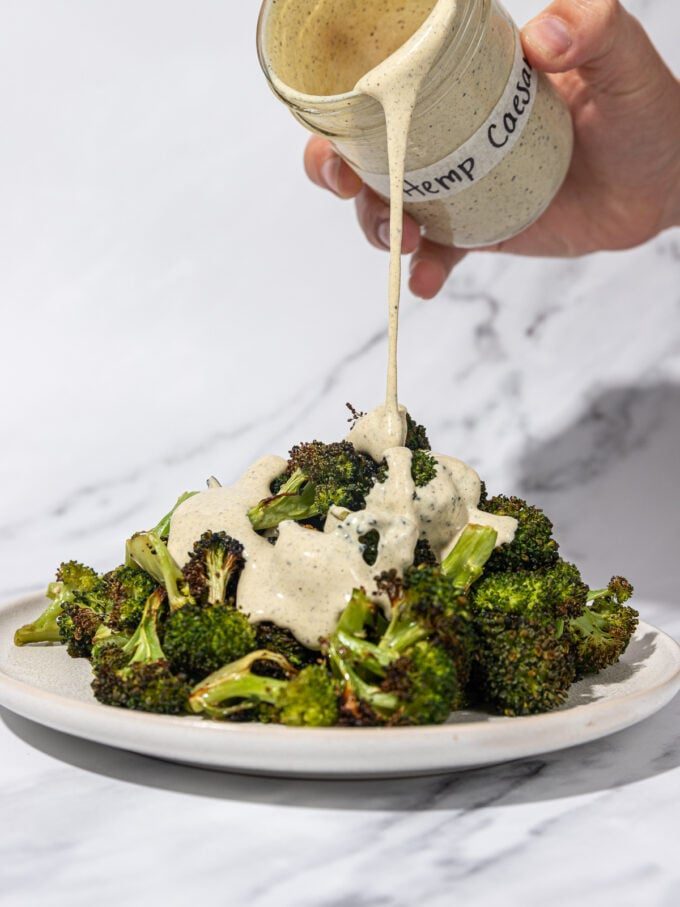hand pouring Caesar dressing over a plate of roasted broccoli 