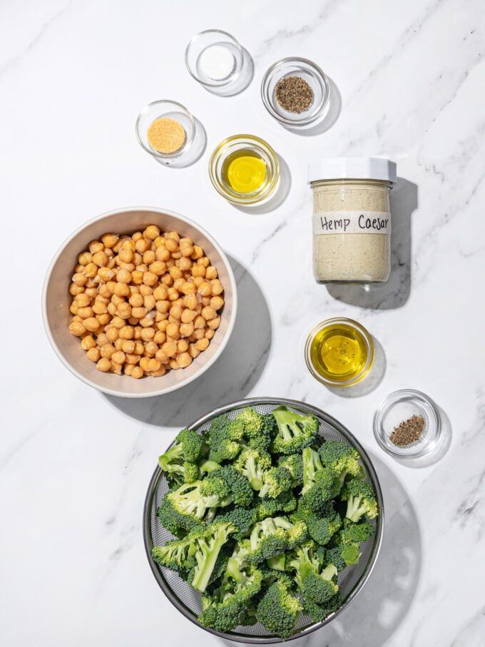 bowls of broccoli, chickpeas, oil, spices, and a jar of hemp caesar dressing, flat laid on a marble background