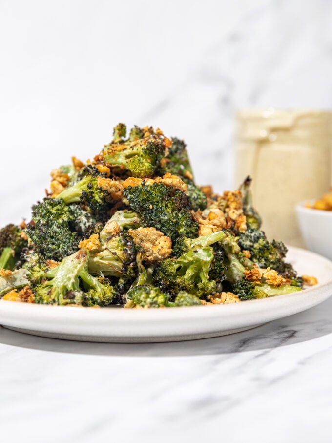roasted broccoli with crumbled chickpeas piled high on a white plate with a glass jar of dressing and white bowl of chickpeas in the background