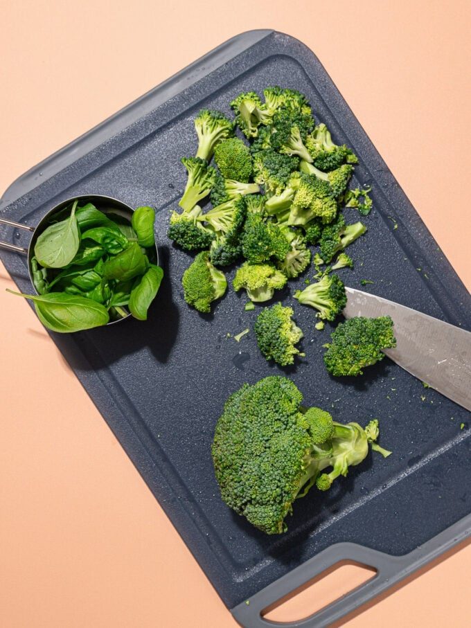 chopped broccoli with knife on black cutting board with a measuring cup full of basil leaves beside it