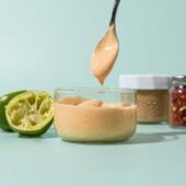 hand holding spoon of cashew dressing over glass jar of dressing, with spent limes, and jars of cashew butter and chile flakes beside it