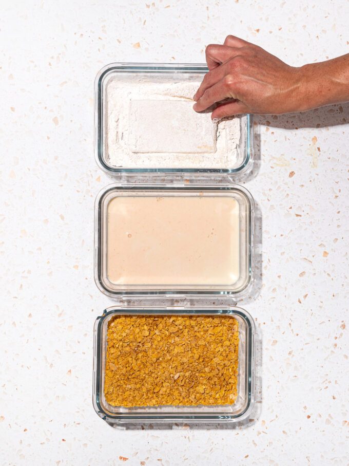 hand dipping tofu into dish of cornstarch with dishes of milk wash and cornflakes beside it